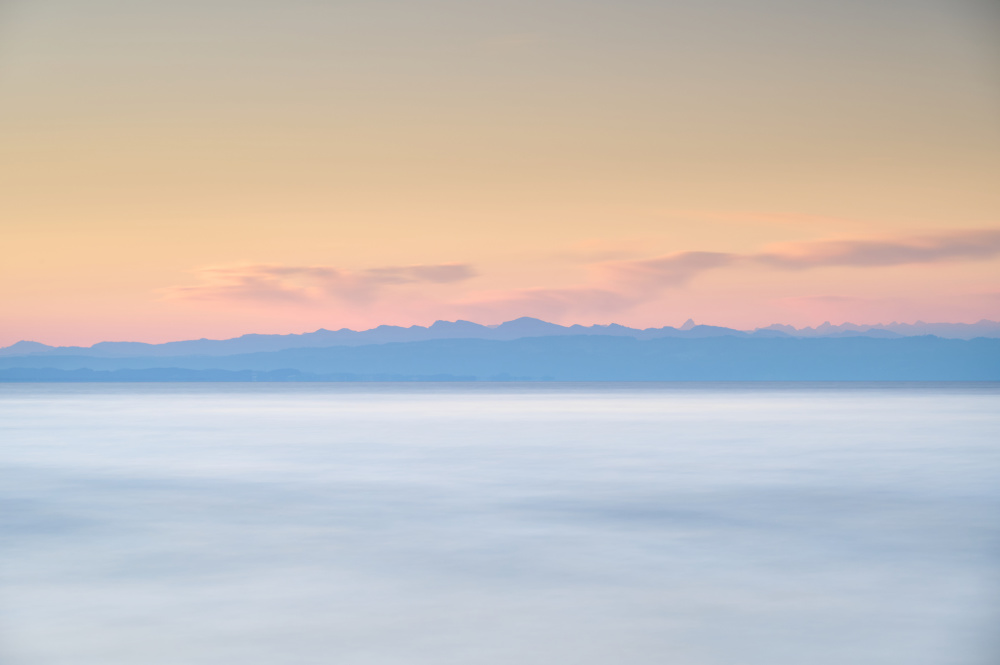 Das Bild zeigt Berge der Alpen hinter dem Bodensee in schöner Lichtstimmung am frühen Morgen. Langzeitbelichtung.